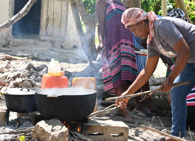 Two large pots sit on cinderblocks and rocks above an open fire. A woman in head wrap, T-shirt, and jeans feeds sticks into the fire.