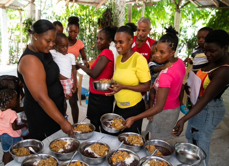 A dozen Haitian adults and small children in bright colors gather around a table, smiling. The mothers reach for metal bowls full of cooked food.