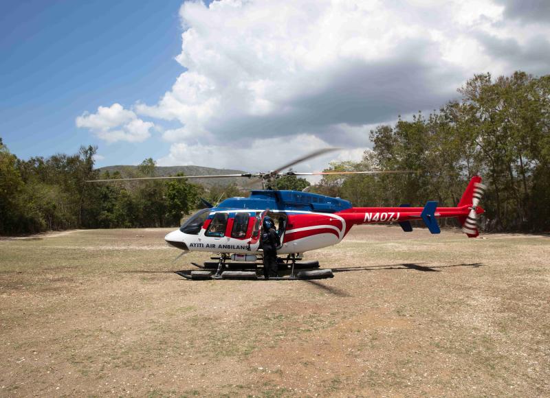 A red, white, and blue helicopter marked Ayiti Air Anbilans, touched down on a dry dirt field under blue skies.