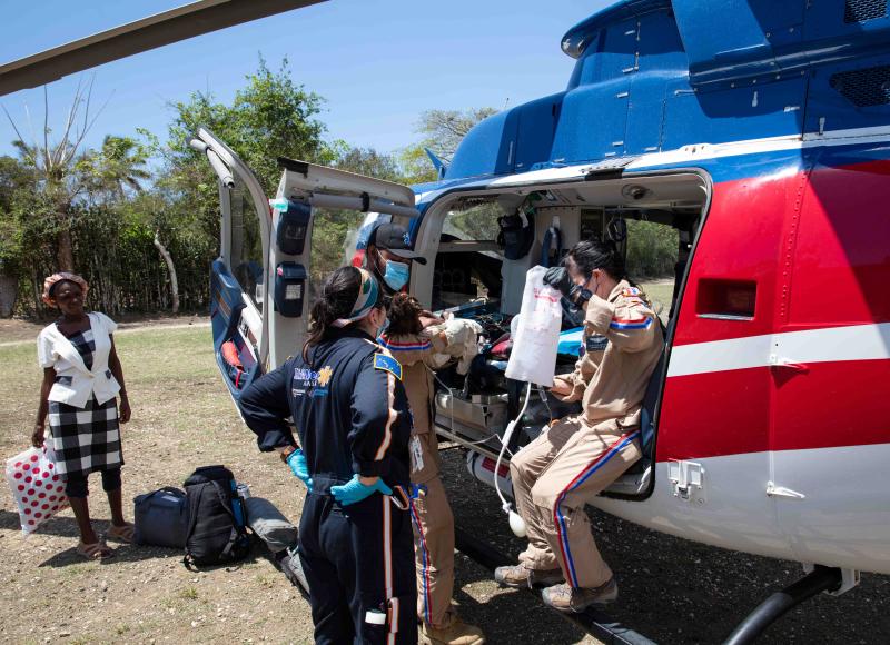 Two women and one man, all in uniforms, gloves, and face masks, lift a baby and IV out of the helicopter. A woman in a dress stands to one side.