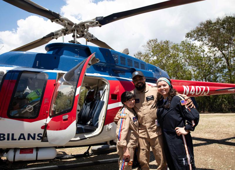 The air ambulance team poses, smiling, in front of their helicopter. A Black man in the middle is flanked by 2 light-skinned women with dark hair.
