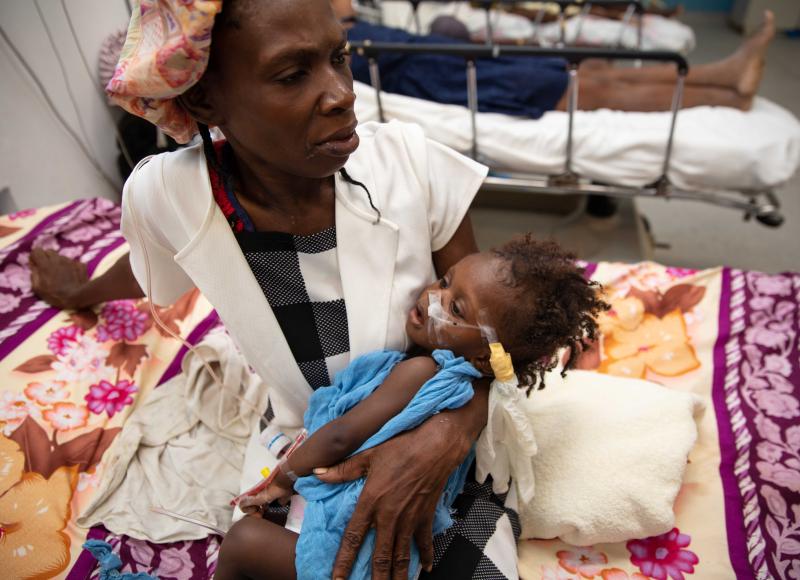 A Haitian woman in church clothes sits on a hospital bed, holding her sick baby on her lap. Other patients can be seen behind them.