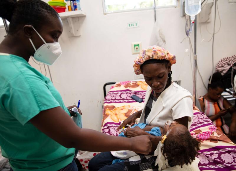 A female clinician in scrubs and hospital mask examines a baby held in his worried mother’s arms. All are Haitian.