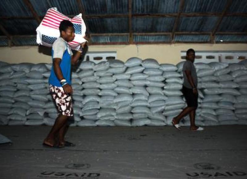 A man carries a huge bag of rice on his shoulder; behind him, there are countless stacked bags of rice.