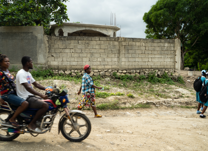 Edmonde, a pregnant woman, walks on a dirt path behind two schoolgirls. A man and woman on a motorcycle pass by on her right.