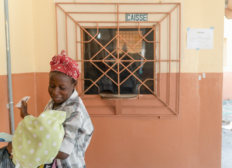 A Haitian woman looks adoringly at her swaddled baby as she walks away from a pink and peach-colored building.