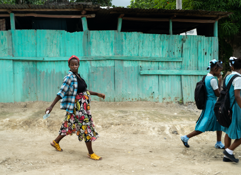 Edmonde, a pregnant woman, walks behind two school girls on a dirt path with a blue house on her left side.
