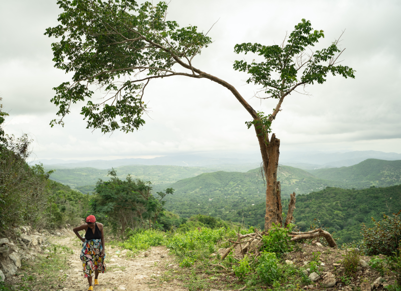 Edmonde, a pregnant woman, walks on a dirt path while look down. A large tree stands to her left and there are green mountains in the background.