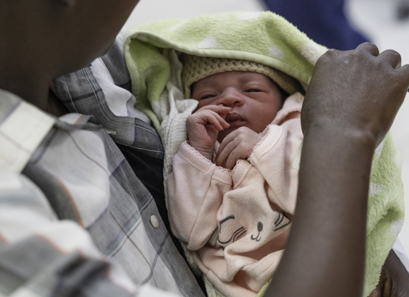 A Haitian woman holds a her newborn baby. The baby wears a light pink outfit and is swaddled in a light green blanket.