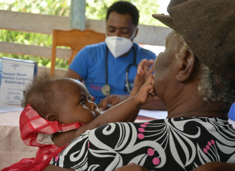 A woman holds a baby while recieving care at an earthquake mobile clinic. The baby reaches out and touches the woman's face while a doctor looks on in the background.