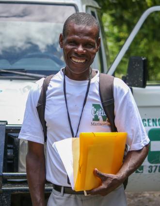 A Haitian man wearing a white T-shirt, backpack, and lanyard stands in front of a hospital vehicle, grinning. He holds papers and manila envelopes in his left hand.