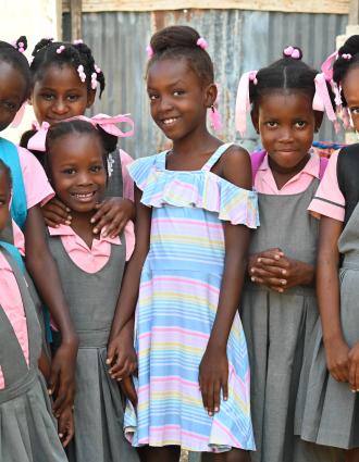 A girl in blue & white dress smiles shyly and happily. She is surrounded by 7 other smiling girls in gray & pink school uniforms.