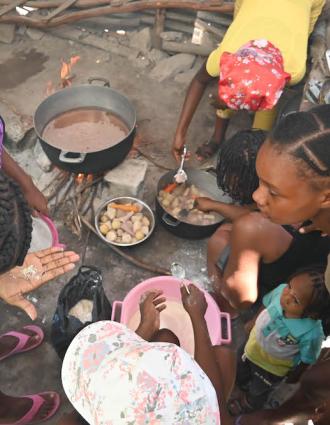 A group of Haitian women crowd around pots and pans to cook a mid-day meal for their children.