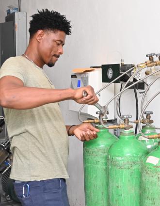 A Haitian man uses a large wrench to remove a green oxygen cylinder from an array of cylinders and hoses. On his left are blue compressors and a large black tank.