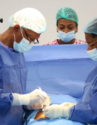 Two Haitian surgeons in full PPE, one male and one female, begin operating on a patient. Another clinician observes from behind a blue drape.