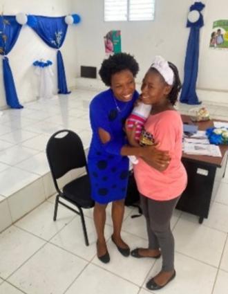 2 Haitian women embrace, smiling, in a white tiled room decorated in blue. One wears a blue dress and the other wears pink and holds an infant.