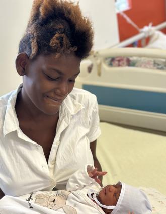 A young Haitian woman in a white blouse smiles down at the infant she holds in her arms.