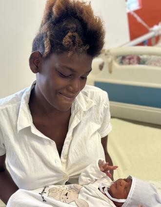 A young Haitian woman in a white blouse smiles down at the infant she holds in her arms.