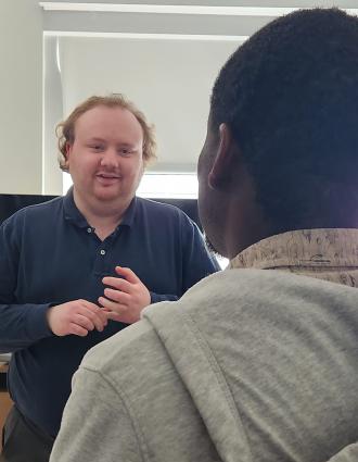 In a neutral office space, a white man with reddish hair speaks to a Haitian man in a gray hooded sweatshirt.