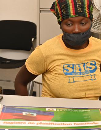 A woman in gold T-shirt, black/green/gold/red head scarf, and face mask sits facing a desk with office supplies and an oversize book with an image of the Haitian flag on its cover.