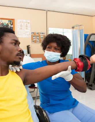 A Haitian man wearing a yellow tank top does arm strengthening exercises with a Haitian physical therapist, who wears blue scrubs and a medical mask.