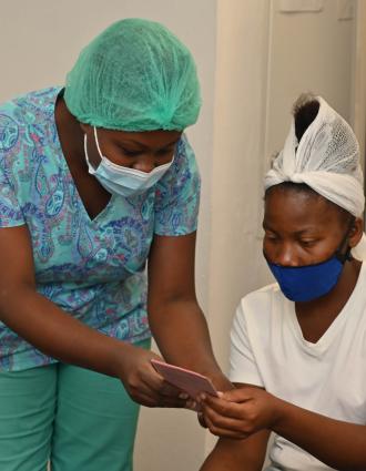 A Haitian woman in bright aqua scrubs and face mask smiles as she hands a pink card to another Haitian woman wearing a white shirt and head wrap and blue face mask.