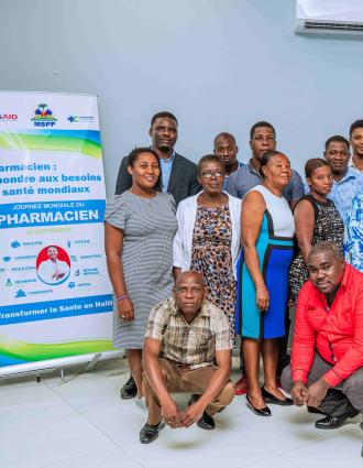 22 Haitian men and women, in business casual attire, pose for a group photo next to a World Pharmacists Day banner, in French, with USAID, MSPP, and HEI logos.