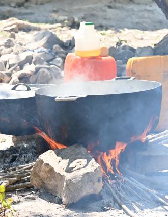 Two large pots sit on cinderblocks and rocks above an open fire in a sunny yard. One pot is covered, and the other has steam rising from it.