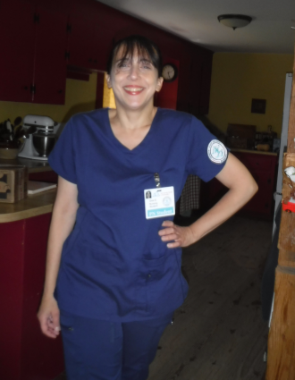 A light skinned woman stands in a wood-framed doorway dressed in dark blue medical scrubs. She has one hand on her hip and smiles at the camera.