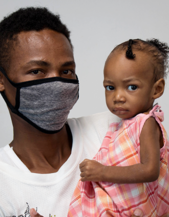 A Haitian man wearing a grey cloth face mask holds his baby daughter in his arms. She is wearing a pink plaid dress and has her hair up.