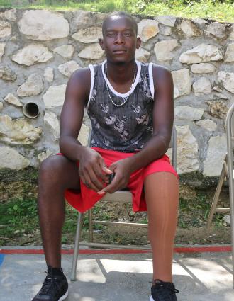 A tall Haitian man sits in a metal folding chair looks at the camera with his hands in his lap. His left leg is a prosthetic.