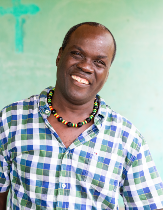 A Haitian man wearing a beaded necklace and a checkered shirt stands in front of a green wall and smiles.