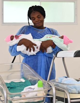 In a hospital room, a Haitian woman in a blue surgical gown holds two newborns with pink hats. A third newborn rests in a bassinet. 