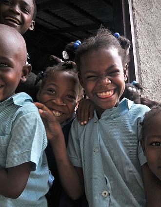 A group of five Haitian school children laugh and smile. They are wearing blue checked shirts.