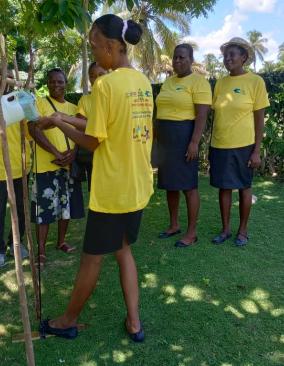 A woman wearing a bright yellow t-shirt demonstrates how to use a handwashing station. A group of five women in the same yellow t-shirt stand around her and observe.