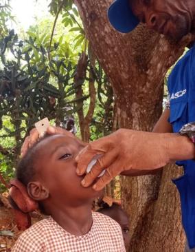 In the Haitian countryside, a man wearing a bright blue cap and a vest with official logos holds a small cup up to a young boy’s mouth.