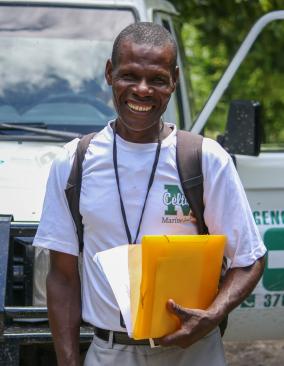 A Haitian man wearing a white T-shirt, backpack, and lanyard stands in front of a hospital vehicle, grinning. He holds papers and manila envelopes in his left hand.
