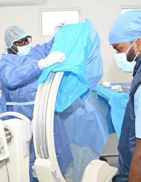In a white operating room, two Haitian men in full PPE prepare for surgery. One arranges blue drapes over a C-arm device and the other checks a monitor.