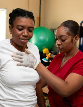 Two Haitian women, seen from the waist up, stand in a brightly-lit rehab center. One, in a red scrub top, presses a hand to the other woman’s upper chest, supporting her.