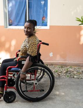 A Haitian woman in a wheelchair looks tot he side as she rolls down a gravel path in front of a peach and cream-colored building.