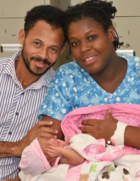 A light-skinned man with neatly-trimmed facial hair and a darker-skinned woman holding an infant in a pink blanket lean into each other, smiling happily.