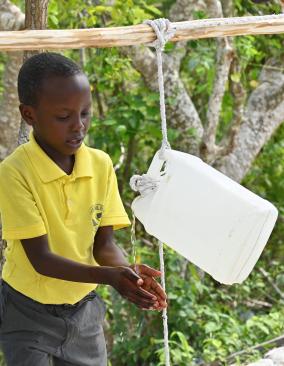 A young boy and girl in yellow school polo shirts wash their hands using a Tippy Tap device.