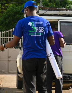 In a courtyard, men unload cardboard boxes from a 4WD vehicle. 1 wears a bright blue cap and T-shirt with HEI/SBH logo.