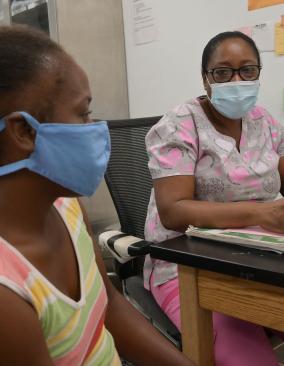 Two Haitian women wearing face masks sit in a medical office. One, in pink scrubs, looks warmly at the camera from behind a desk, while the other woman, in striped top, is seen in profile.