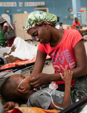 A woman in a red T-shirt that says “Love” sits with a young boy on a hospital bed, gently supporting his head. He curls into her, holding her arm.