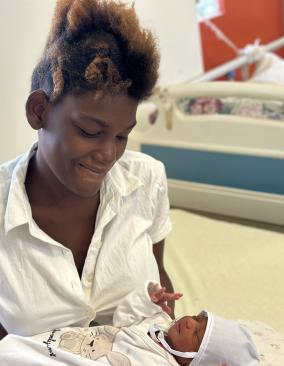 A young Haitian woman in a white blouse smiles down at the infant she holds in her arms.