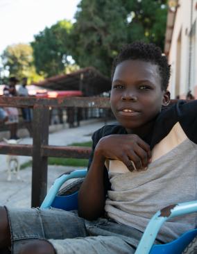 A Haitian teen sits in a bright blue wheelchair. His upper body is turned to face the camera. He is smiling.