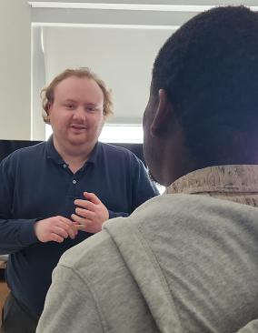 In a neutral office space, a white man with reddish hair speaks to a Haitian man in a gray hooded sweatshirt.