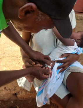 A male community health worker vaccinates a baby, who sits in his mother's lap.