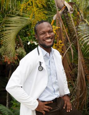 A Haitian male doctor stands in front of palm trees and smiles brightly a the camera. He has his hands on his hips. He wears a light blue button-down shirt and a white doctor's coat with a stethescope over his shoulders.